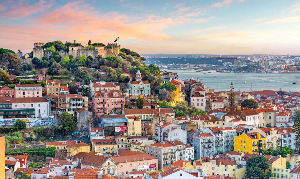 Panoramic view of Lisbon, Portugal, featuring colorful hillside buildings and the historic São Jorge Castle, overlooking the Tagus River at sunset.